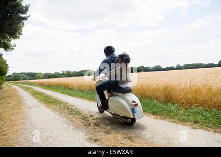 Rear view of mature man and daughter riding motor scooter along dirt track Stock Photo
