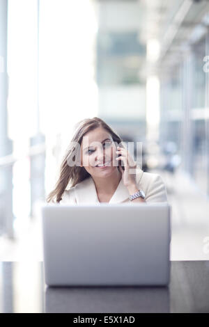Young businesswoman using laptop and chatting on smartphone in conference centre Stock Photo