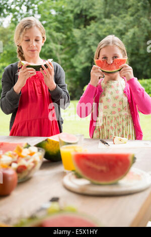 Two sisters at patio table eating and holding up watermelon slice Stock Photo