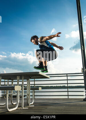 Young man jumping backwards from picnic table on lake pier Stock Photo