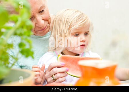 Senior woman and toddler great granddaughter eating melon slice at garden table Stock Photo