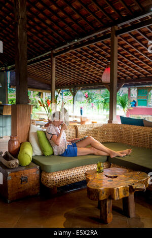 Woman relaxing with coffee and digital tablet in holiday apartment sitting room, Ubud, Bali, Indonesia Stock Photo