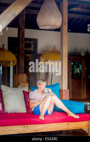 Woman relaxing with coffee and digital tablet in holiday apartment sitting room, Ubud, Bali, Indonesia Stock Photo