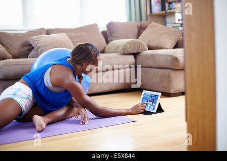 Young woman exercising on sitting room floor whilst using touchscreen on digital tablet Stock Photo