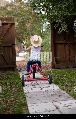 Four year old girl riding her tricycle out of garden gate Stock Photo