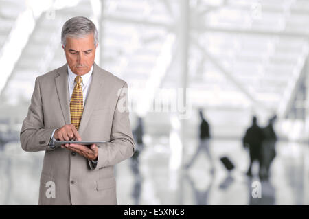 Closeup of a mature businessman using his tablet computer in an airport concourse. The man is looking at the device with blurred Stock Photo