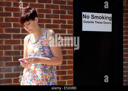 young woman with cigarette and mobile phone in front of a no smoking sign Stock Photo