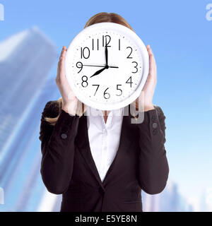 Business woman wearing formal suit and holding big clock on face, standing outdoors on office building background Stock Photo