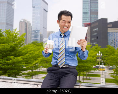 asian businessman looking at tablet holding coffee cup smiling. Stock Photo