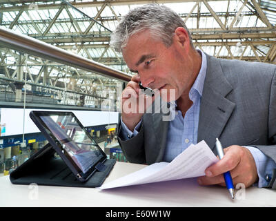 Mature businessman seated at cafe table on railway concourse looking at his tablet computer screen and using his smartphone Stock Photo