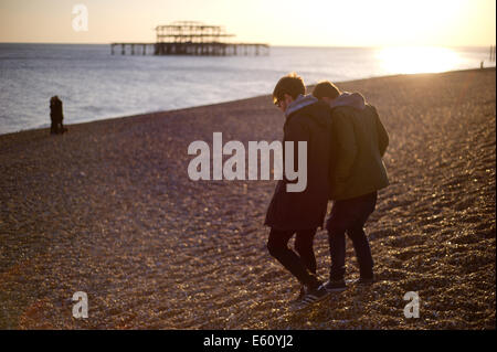 Two people walk along Brighton beach at sunset. The burned down West Pier can be seen in the background. Stock Photo