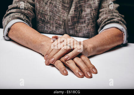 Detail of old woman's hands resting on grey surface. Senior female's hand on top of another while sitting at a table. Stock Photo