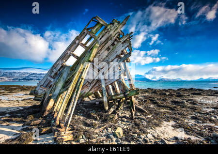 The wooden skeleton of a ship wrecked on a stony beach against a blue clouded sky with snow covered mountains behind Stock Photo
