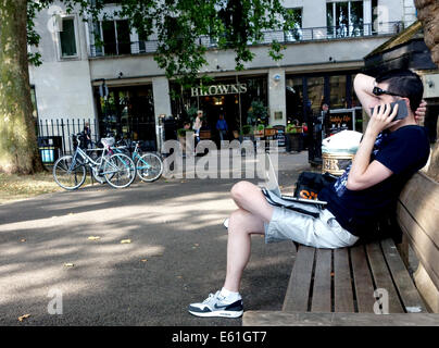 Man on park bench using laptop computer and smartphone, London Stock Photo