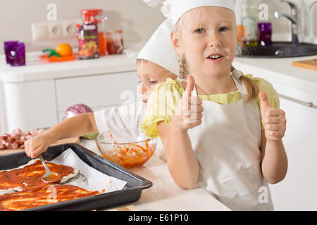 Little girl giving a thumbs up as she bakes pizza with her young brother who is busy spreading tomato paste onto the bases in the baking tray. Stock Photo