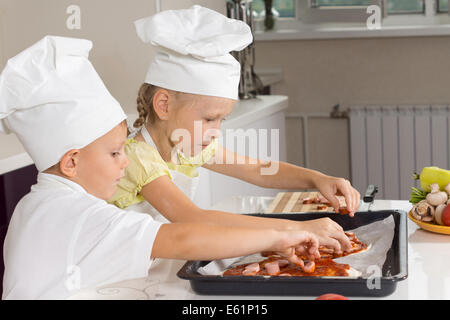 Young girl and boy in white chefs uniforms loading ingredients onto the homemade pizza bases they have just made before placing Stock Photo