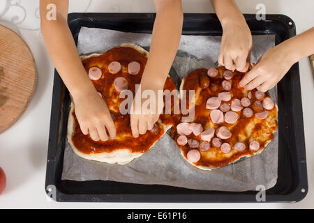 Two small children making themselves a traditional Italian pizza for lunch placing sliced sausage onto the tomato paste and pastry for the base, closeup view of their hands. Stock Photo