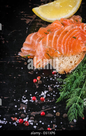 Bread with sliced salted salmon, served on black wooden table with lemon, sea salt and peppers. Stock Photo