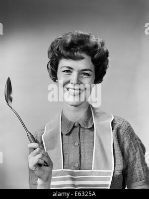 1950s 1960s PORTRAIT SMILING WOMAN HOUSEWIFE WEARING APRON HOLDING LARGE SPOON Stock Photo