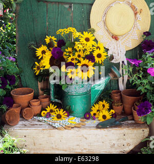 BOUQUET YELLOW FLOWERS IN GREEN WATERING CAN HAT AND GARDENING TOOLS ON WOODEN BENCH Stock Photo