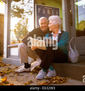 1990s COUPLE SITTING ON STEP OUTSIDE TRAVEL AGENCY READING BROCHURE Stock Photo