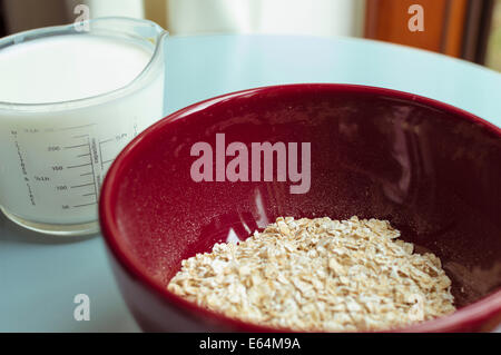 porridge oats in a red bowl; milk-filled measuring jug in the background on a blue table-top. Stock Photo