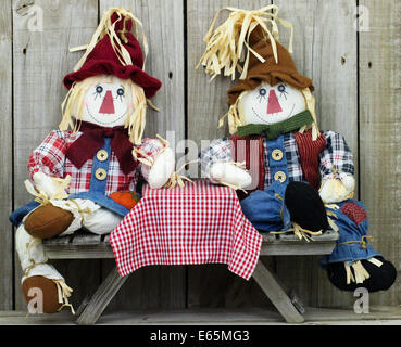 Boy and girl scarecrows sitting on picnic table with red checkered tablecloth by wood fence Stock Photo