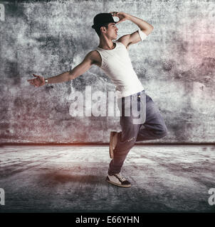 Hip young man in a tank top and hat doing a dance routine posing on one leg in a grunge concrete room or stage, profile full Stock Photo