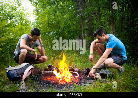 Two young men camping in the mountains crouching alongside a burning campfire with rucksack staring thoughtfully at the fire Stock Photo