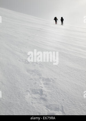 two men with heavy backpacks crossing a snowfield on snowshoes in  winter mountains with tracks in forground Stock Photo