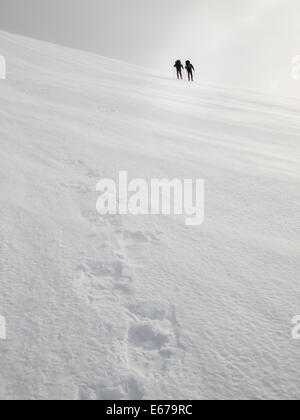 two men with heavy backpacks crossing a snowfield on snowshoes in  winter mountains with tracks in foreground Stock Photo
