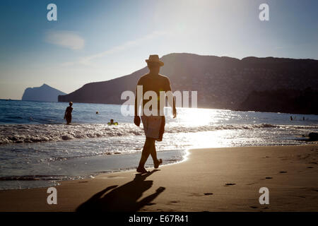 Man wearing a straw trilby style hat and shorts walks along a sandy beach towards the late afternoon sun. Stock Photo