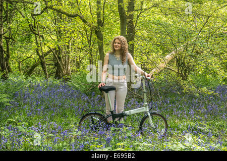 A pretty teenager, standing smiling next to her bike amongst bluebells in the woods at Banstead, Surrey, England, UK. Stock Photo