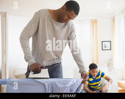 Father ironing shirt while son plays video games Stock Photo