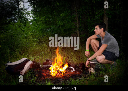 Young man camping in the mountains crouching alongside a burning campfire with his rucksack staring thoughtfully into the Stock Photo