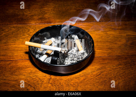 A burning cigar in a classic black ashtray streaming smoking in a dark, moody setting. Stock Photo