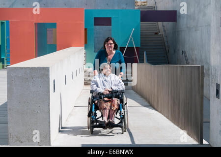An elderly lady is pushed up a concrete disabled access ramp in a wheelchair by her carer or assistant on a sunny day. Stock Photo