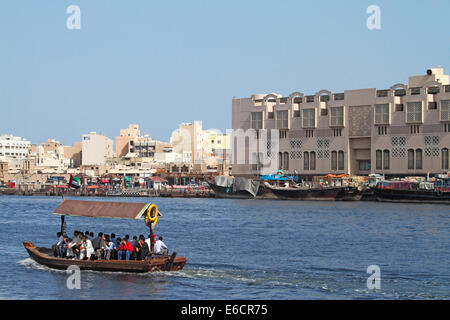 Wooden boat, traditional abra used as small public transport ferry, with passengers on Dubai Creek, with waterfront buildings & dhows in city of Dubai Stock Photo