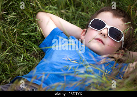 young Caucasian boy in blue shirt with white sunglass  lying on a big green grass, relaxing. Stock Photo