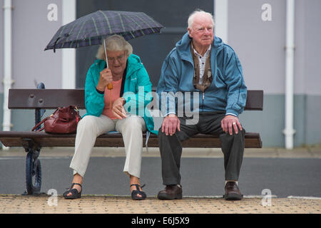 Aberystwyth, Wales, UK. 25th August, 2014. A senior couple sitting on a seaside bench in the wet, cold and overcast weather at Aberystwyth on the west Wales coast on August Bank Holiday Monday 2014. The forecast is for persistent heavy rain to hit the wet coast throughout the day photo Credit:  keith morris/Alamy Live News Stock Photo