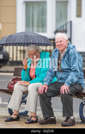 Aberystwyth, Wales, UK. 25th August, 2014. Looking cheerful, a senior couple sit on a seaside bench in the wet, cold and overcast weather at Aberystwyth on the west Wales coast on August Bank Holiday Monday 2014. The forecast is for persistent heavy rain to hit the wet coast throughout the day photo Credit:  keith morris/Alamy Live News Stock Photo