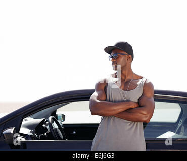 Image of young african man standing by his car. Muscular male model wearing sunglasses and cap outdoors. Stock Photo