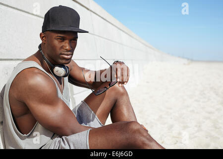 Image of muscular young african man sitting on a beach. Male model wearing a cap and headphones holding sunglasses looking away. Stock Photo
