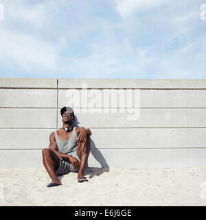 Image of young man relaxing on the beach. African male model leaning on a wall. Guy wearing cap and sunglasses resting on beach. Stock Photo