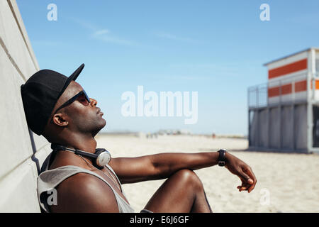 Side view of relaxed African man leaning to a wall at beach sunbathing. Black guy wearing sunglasses and cap relaxing. Stock Photo
