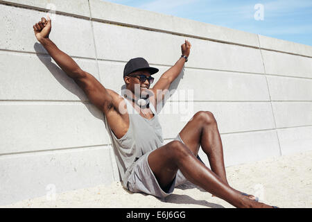 Muscular young man sitting by a wall stretching his hands smiling. African male model on beach. Stock Photo