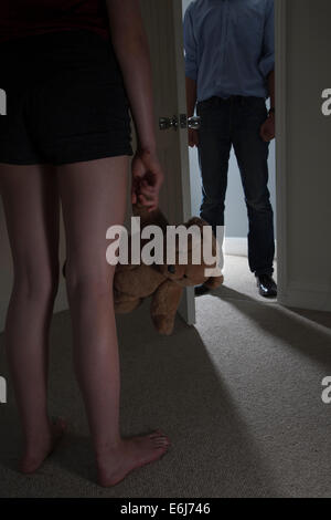 Anonymous man entering a dark room, a young girl standing in the foreground looking towards the man holding a teddy-bear. Close. Stock Photo