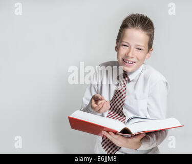 The nice laughing boy teenager in a white shirt and a tie reads a big red funny book on a gray horizontal background Stock Photo