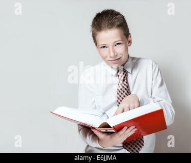 The nice smiling school boy in a white shirt and a tie reads a big red book on a gray background Stock Photo