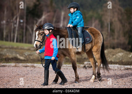 Girl leading a pony with a young child riding it, dun, with a bridle, Tyrol, Austria Stock Photo